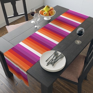 Puede incluir: Un camino de mesa colorido con un patrón de rayas en tonos naranja, rojo, rosa y blanco. El camino de mesa está colocado sobre una mesa de madera oscura, con un cuenco de manzanas, copas de vino y un plato con cubiertos.