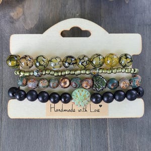 Puede incluir: Un conjunto de pulseras de cuentas exhibidas en una tarjeta de madera. Las pulseras presentan una variedad de cuentas en tonos verdes, marrones y negros. Una pulsera tiene una cuenta verde decorativa. La tarjeta tiene el texto "Handmade with Love."