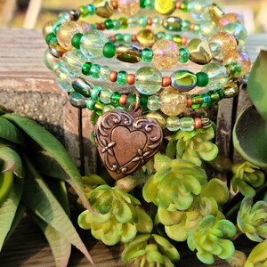 Op de afbeelding: Een stapel armbanden met kralen in groen, goud en helder. De armbanden hebben verschillende kralenvormen en -maten, met een koperen hartbedel. De armbanden worden gepresenteerd op een houten oppervlak met groen gebladerte.