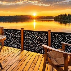 May include: A black metal deck railing panel featuring a laser-cut zebra stripe pattern installed between wooden posts on a wooden deck overlooking a lake at sunset with a wooden chair and a round table with a mug visible in the foreground.