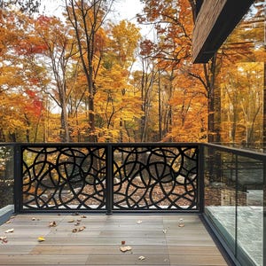 May include: Black metal railing with a decorative, circular pattern on a wooden deck. The railing is in front of a forest with trees displaying vibrant autumn colors of yellow, orange, and red. A glass panel is to the right of the railing.