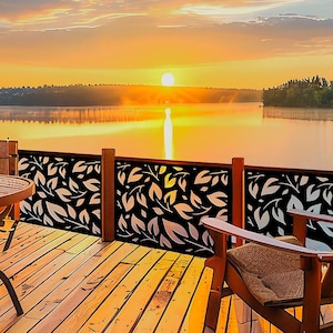 May include: A black metal deck railing panel featuring a repeating laser-cut leaf pattern. The panel is installed on a wooden deck with a table and chair, overlooking a calm lake during a bright orange sunset with trees on the horizon.