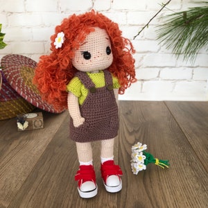 May include: A crocheted doll with red curly hair, wearing a green and brown outfit and red shoes. The doll is holding a bouquet of white crocheted flowers.