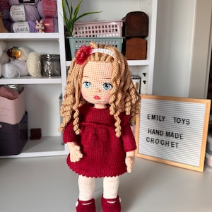 Puede incluir: Una muñeca de crochet con el pelo largo y rizado rubio, que lleva un vestido rojo, medias blancas y zapatos rojos. La muñeca está de pie frente a una estantería blanca con ovillos de lana y un cartel que dice "Emily Toys Hand Made Crochet".