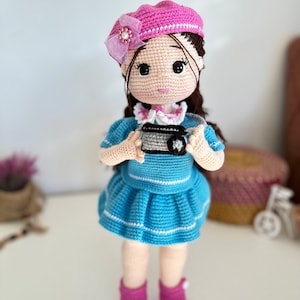 May include: A crocheted doll wearing a pink beret, a blue and white striped dress, and pink shoes. The doll is holding a small camera.