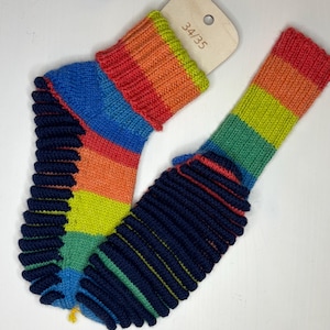 Op de afbeelding: Een paar kleurrijke gebreide sokken met een regenboogstreepjespatroon aan de bovenkant en marineblauwe geribbelde details op de voet. Een houten label met de maat "34/35" is bevestigd. De sokken zijn ontworpen voor comfort en stijl.