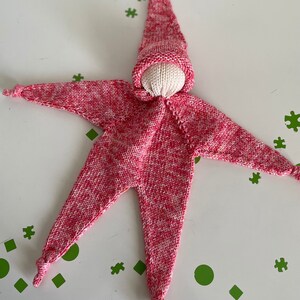 Puede incluir: Una muñeca de punto rosa y blanco con un gorro puntiagudo. La muñeca tiene un cuerpo en forma de estrella y está atada en las piernas y los brazos.