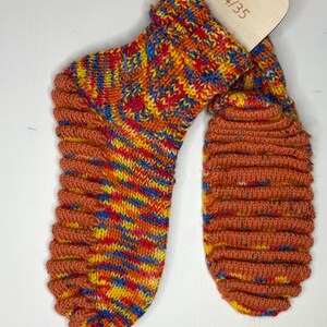 Puede incluir: Un par de calcetines de punto coloridos con puño y puntera acanalados. Los calcetines presentan una mezcla de hilos rojos, amarillos, azules y naranjas. Una etiqueta de madera con la talla 34/35 está adjunta.