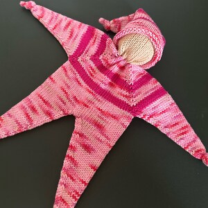 May include: A knitted star-shaped baby toy in pink and red hues. The toy features a light beige head and a matching pointed hat. The star's arms and legs are long and tapered, with knotted ends.
