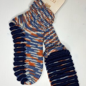Puede incluir: Un par de calcetines de punto con un patrón de rayas azules, naranjas y blancas. Los calcetines tienen un puño y una suela de color azul oscuro. Una etiqueta de madera indica la talla 32/33.