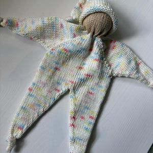 May include: A handmade, star-shaped doll with a beige head and a white knitted body. The doll's body is speckled with pastel colors, including pink, blue, and yellow. It has a hood and long legs, and the arms are outstretched.