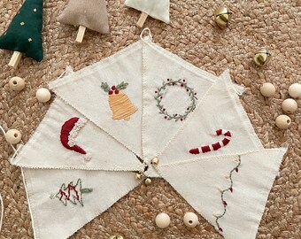 May include: A set of six white fabric triangle flags with different Christmas-themed embroidery designs, including a bell, a wreath, a candy cane, a Christmas tree, a Santa hat, and a string of lights. The flags are strung together with white twine and wooden beads.