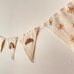 May include: A white fabric banner with embroidered designs. The banner features a cloud, a rainbow, and stars. The banner is strung up on a wall.