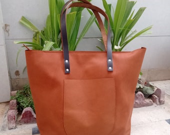 May include: A brown leather tote bag with a large front pocket and black leather handles. The bag has a zipper closure and is sitting on a tiled floor.