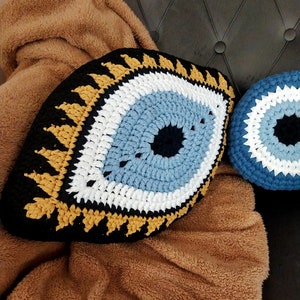 May include: Two crocheted evil eye pillows, one large and one small, with blue, white, black, and gold yarn. The pillows are on a brown fuzzy blanket.