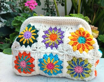 May include: A cream-colored crochet clutch with a floral pattern. The flowers are in various colors, including orange, yellow, blue, green, and purple. The clutch is made of a soft, textured yarn and has a rectangular shape.