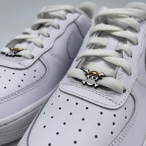 Peut inclure: Des baskets en cuir blanc avec des lacets blancs et un charme en or et noir en forme de crâne et d'os croisés sur les lacets. Le charme a un chapeau rouge et jaune sur le dessus du crâne.