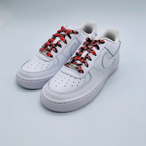 Puede incluir: Zapatillas bajas de cuero blanco con cordones de patr&oacute;n rojo y negro. Los cordones tienen un patr&oacute;n repetido de nubes rojas sobre un fondo negro.