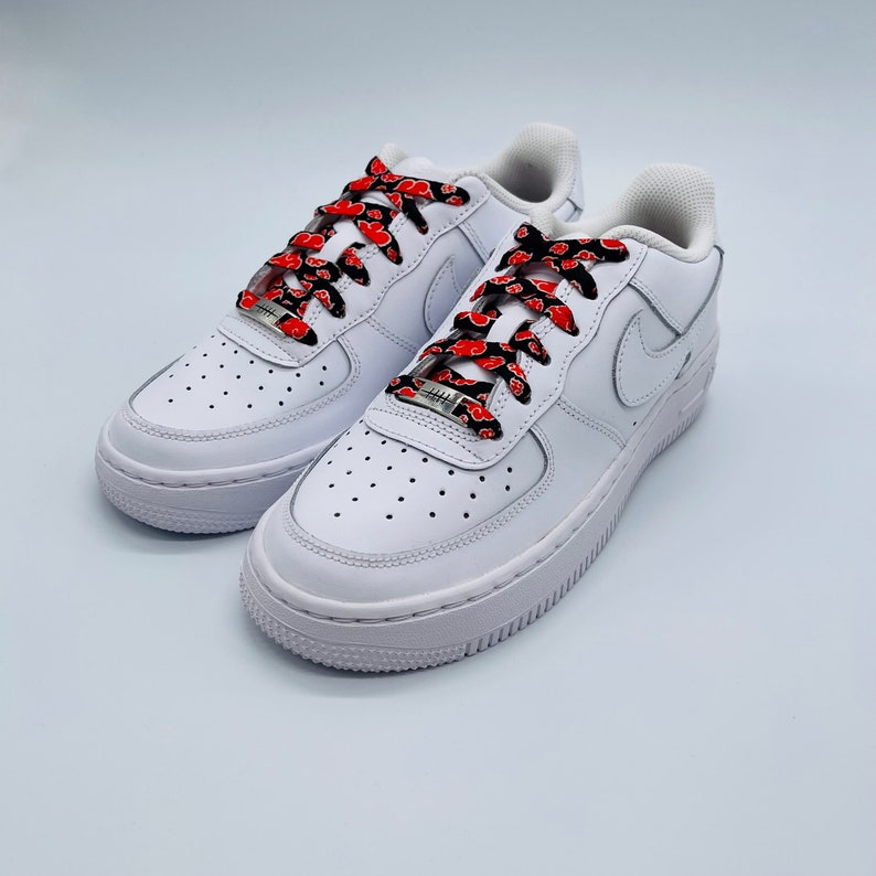 Puede incluir: Zapatillas bajas de cuero blanco con cordones de patr&oacute;n rojo y negro. Los cordones tienen un patr&oacute;n repetido de nubes rojas sobre un fondo negro.