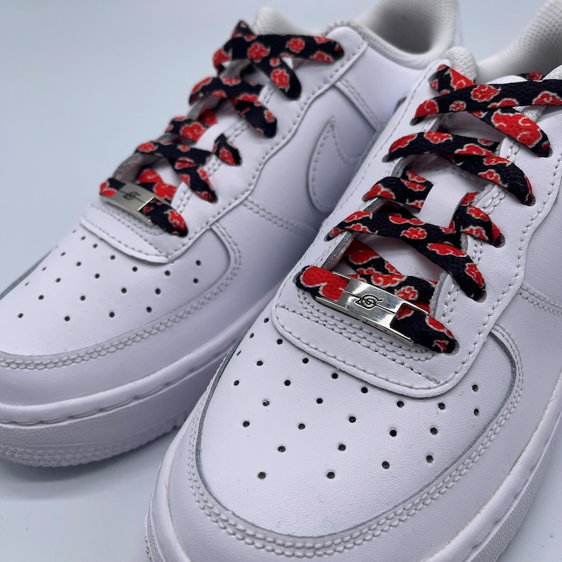 Puede incluir: Zapatillas blancas de cuero con cordones negros y rojos que presentan un patr&oacute;n repetido de nubes rojas. Los cordones est&aacute;n atados con un cierre de cord&oacute;n de metal plateado con un dise&ntilde;o de espiral estilizado.