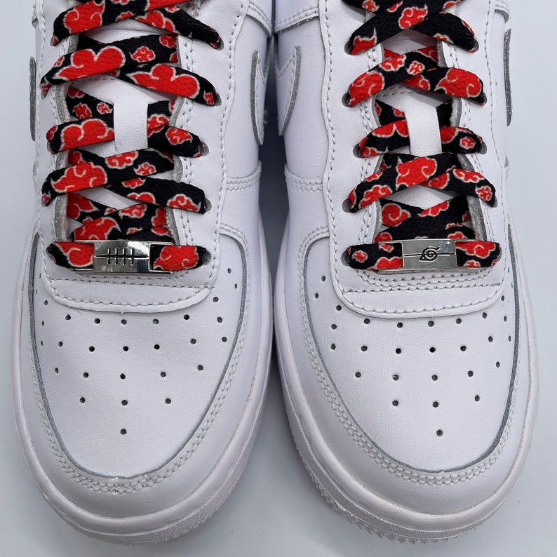 Puede incluir: Zapatillas blancas de cuero con cordones rojos y negros con un patr&oacute;n de nubes. Los cordones tienen aglets de metal plateado con los kanji para "Sharingan" y "Uchiha" en ellos.