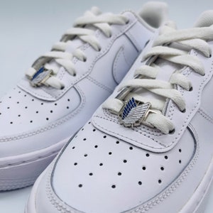 Pode incluir: Um par de tênis brancos com cadarços brancos e pingentes decorativos prateados e azuis. Os pingentes apresentam um design de asa. Os tênis têm um design clássico de baixo perfil com biqueira perfurada.