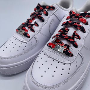 Puede incluir: Zapatillas blancas de cuero con cordones negros y rojos con un patr&oacute;n repetido de nubes rojas. Los cordones est&aacute;n atados con un cierre de metal plateado.