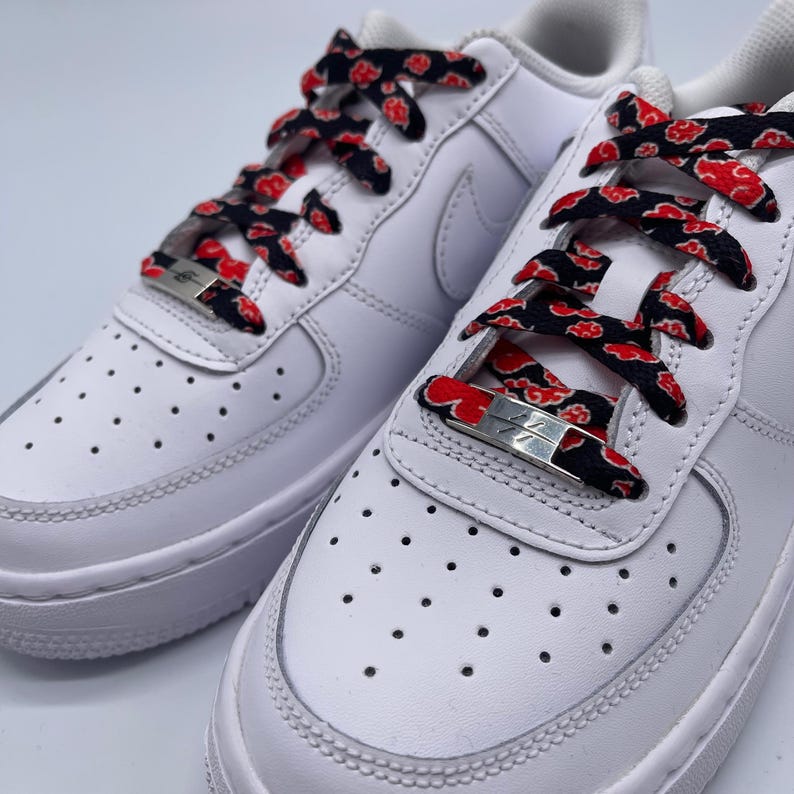 Puede incluir: Zapatillas blancas de cuero con cordones negros y rojos con un patr&oacute;n repetido de nubes rojas. Los cordones est&aacute;n atados con un cierre de metal plateado.