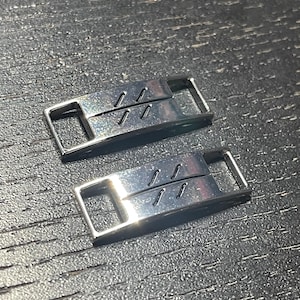 Puede incluir: Dos abalorios de zapatos plateados con un dise&ntilde;o sencillo de tres l&iacute;neas paralelas. Los abalorios son de metal y tienen un acabado brillante.