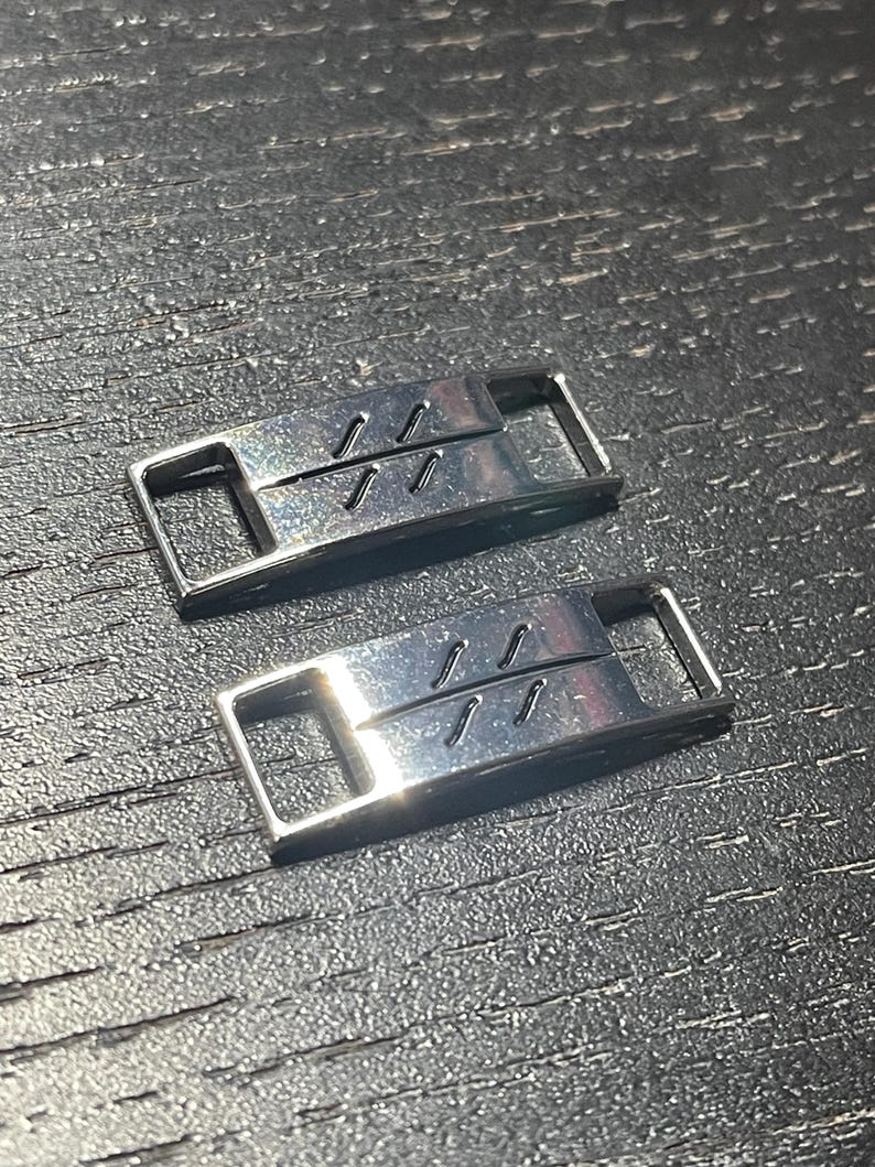Puede incluir: Dos abalorios de zapatos plateados con un dise&ntilde;o sencillo de tres l&iacute;neas paralelas. Los abalorios son de metal y tienen un acabado brillante.