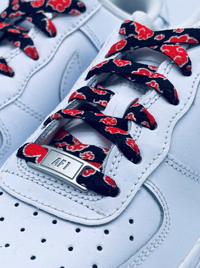 Puede incluir: Zapatillas blancas de cuero con cordones negros y rojos con un patr&oacute;n de nubes rojas. Los cordones tienen un terminal de metal plateado con el texto "AF1" en &eacute;l.