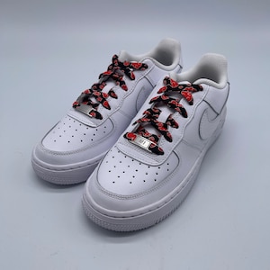 Puede incluir: Zapatillas blancas de ca&ntilde;a baja con cordones de patr&oacute;n rojo y negro. Los cordones est&aacute;n atados con un nudo &uacute;nico y tienen un cierre de cordones de metal plateado con las letras "AF1" grabadas.
