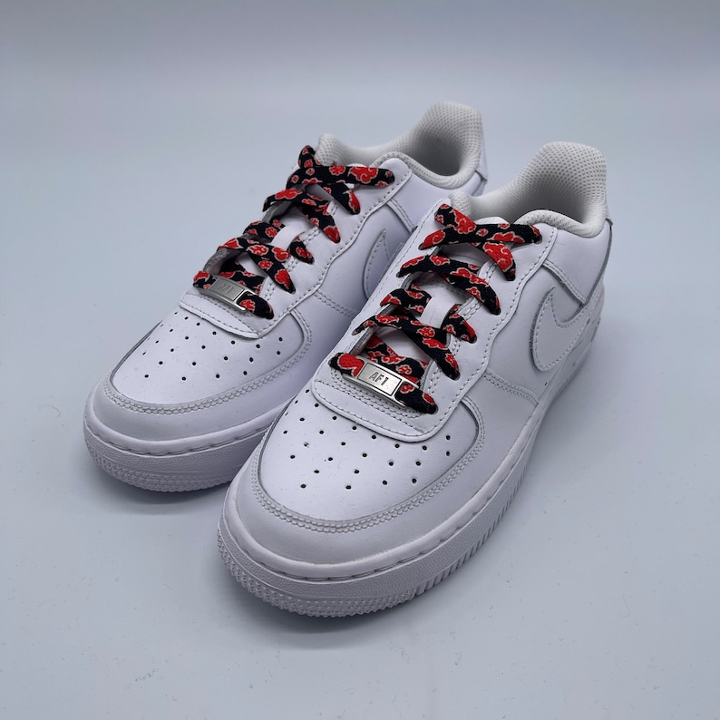 Puede incluir: Zapatillas blancas de ca&ntilde;a baja con cordones de patr&oacute;n rojo y negro. Los cordones est&aacute;n atados con un nudo &uacute;nico y tienen un cierre de cordones de metal plateado con las letras "AF1" grabadas.