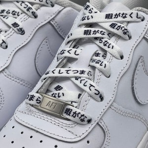 May include: White leather sneakers with white laces featuring Japanese text printed on them. The laces are tied in a criss-cross pattern and the text reads "暇がなくし" and "まらない". The sneakers have a silver metal tag with the letters "AF1" on it.