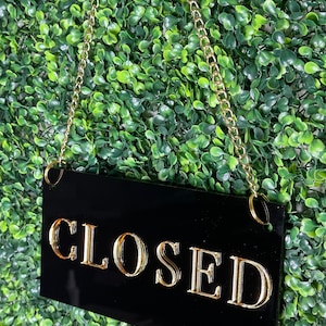 May include: Black sign with gold lettering that reads "CLOSED" hanging from a gold chain against a green backdrop.