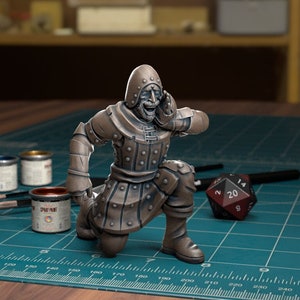 Może przedstawiać: Pomalowana miniatura postaci w kapturze z szerokim uśmiechem, klęcząca z uniesioną ręką do twarzy. Postać jest pomalowana na brązowo i umieszczona na zielonej macie do cięcia z 20-ścienną kostką.