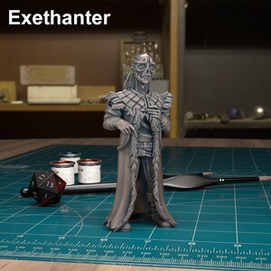 Puede incluir: Una figura miniatura pintada de gris de una figura esquelética que lleva una capa larga y emplumada. La figura está de pie sobre una estera de corte verde con una regla y botes de pintura.