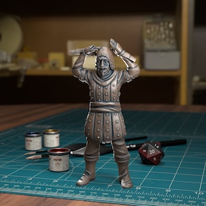 Może przedstawiać: Miniaturowa figurka mężczyzny w zbroi i hełmie, trzymającego miecz nad głową, wykonana z brązu i pomalowana. Figurka stoi na zielonej macie do cięcia z pojemnikami na farbę i kostką do gry.