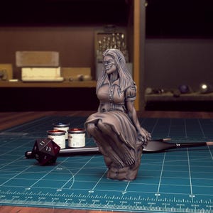 K&ouml;nnte beinhalten: Eine bemalte Miniaturfigur einer weiblichen Figur mit spitzen Ohren, die ein Kleid tr&auml;gt und auf einem Felsen sitzt. Die Figur ist in Bronzefarben bemalt.