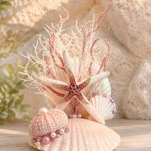 May include: A decorative arrangement featuring a large pale pink scallop shell base, a central starfish adorned with dark pink crystals, a small pink sea urchin test, white pearl beads, and tall, slender, glittery pink and white coral-like branches.