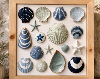 May include: A square light wood frame containing a collection of various seashells and starfish arranged on a white background. The shells are painted in shades of navy blue, light blue, and sage green with white dot and line patterns.