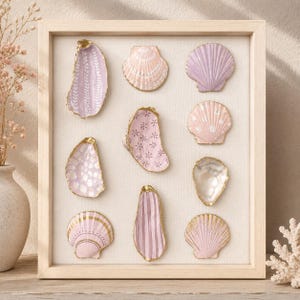 May include: A rectangular light wood frame containing ten decorative seashells arranged in a grid. The shells are painted in shades of pink, lavender, and white with gold-painted edges, featuring patterns like stripes, dots, and floral motifs.