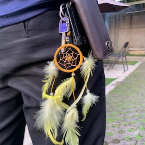 以下が含まれることがあります： キーホルダーに羽根が付いた黄色のドリームキャッチャー。キーホルダーは茶色の革財布に付けられています。