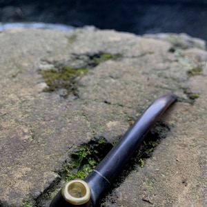May include: A dark brown smoking pipe with a gold-colored rim, resting on a mossy rock. The pipe has a curved stem and a smooth, polished finish. The background is blurred, showing a natural outdoor setting.