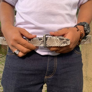 May include: A person wearing a white t-shirt with black paint splatters and the word "OVKL" printed on it. They are wearing dark blue jeans and a snake skin belt with a silver buckle.