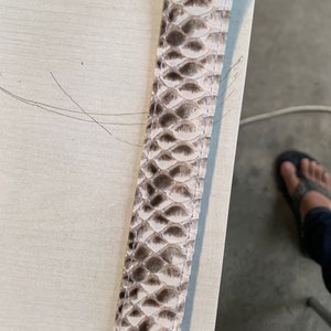 May include: A close-up of a light brown leather strap with a snakeskin pattern. The strap has a white stitching along the edge.
