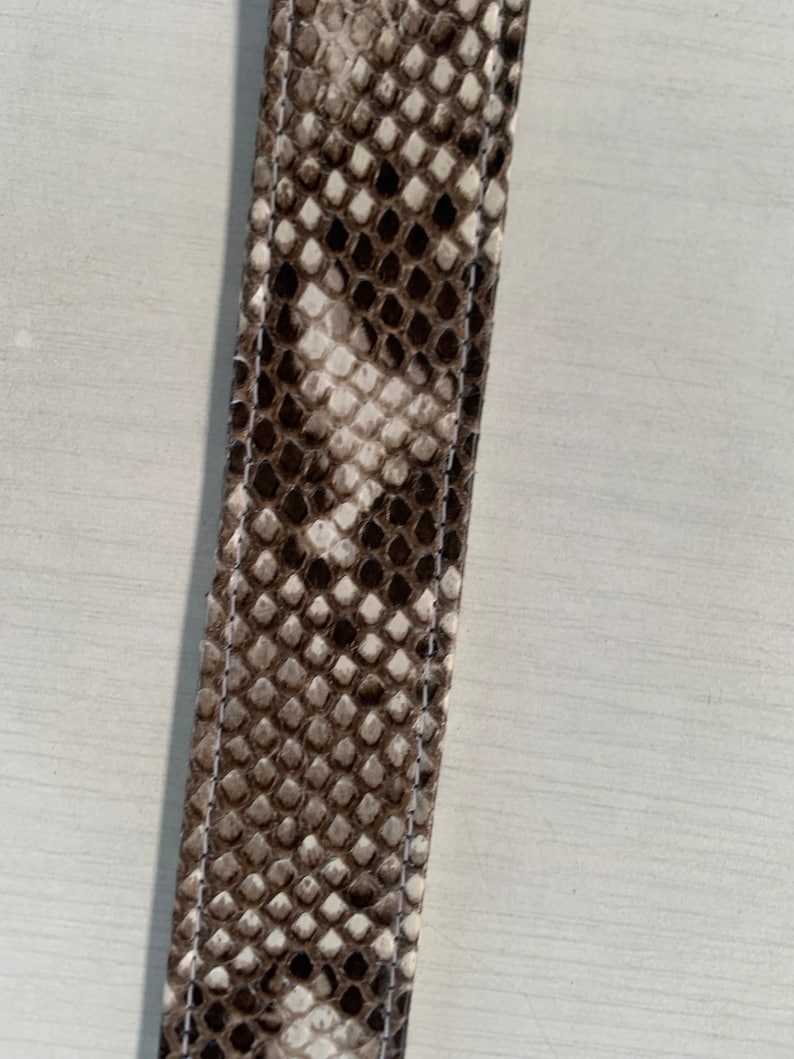 May include: A close-up of a brown and white snakeskin strap with white stitching.