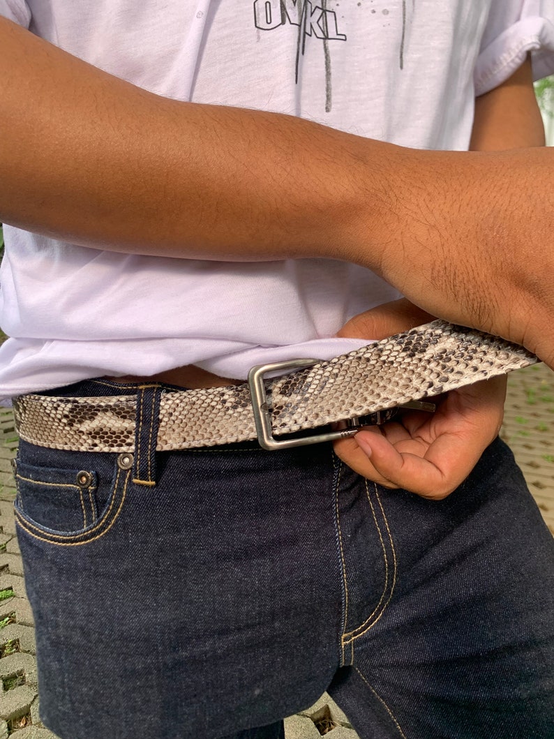 May include: A brown snakeskin belt with a silver buckle. The belt is worn around the waist of a person wearing blue jeans.