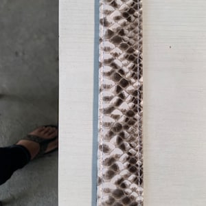 May include: A close-up of a white and brown snakeskin leather strap. The strap has a subtle sheen and a textured surface.