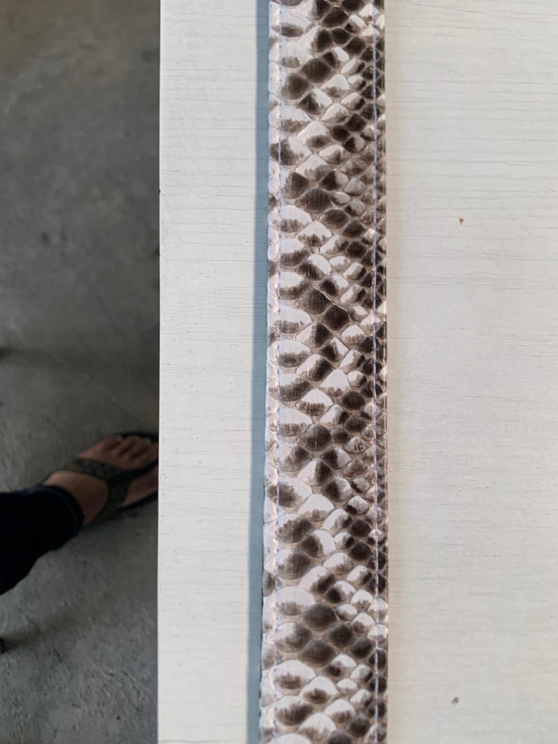 May include: A close-up of a white and brown snakeskin leather strap. The strap has a subtle sheen and a textured surface.